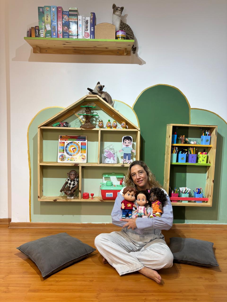 Renata na sala de brincar do Espaço Elo, com estantes em forma de casinha, brinquedos e paredes em arco verde-sálvia