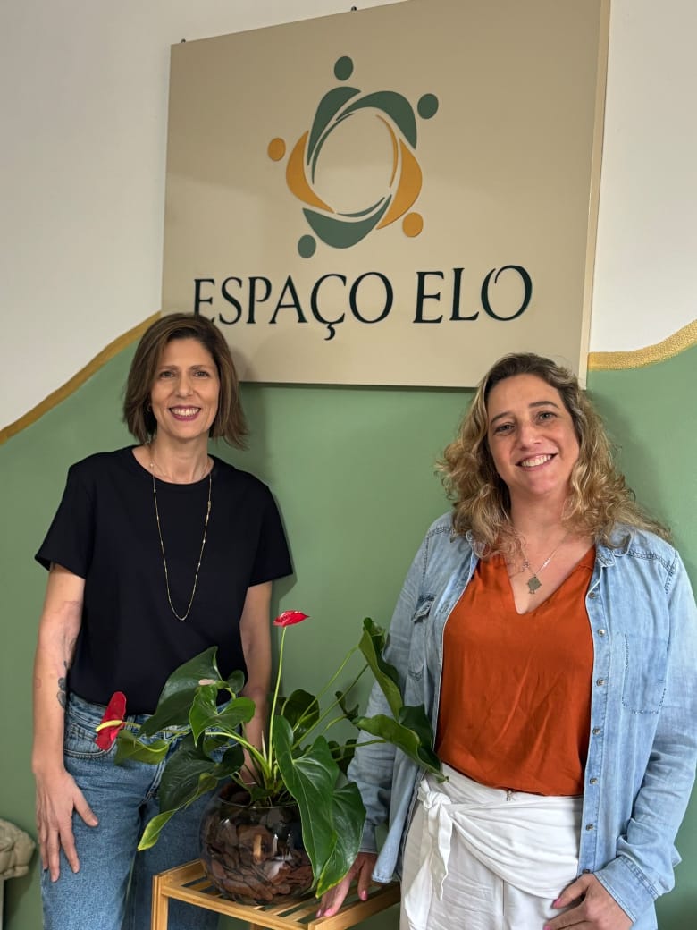 Tatiana e Renata em pé, lado a lado, junto à placa com a logo do Espaço Elo na recepção